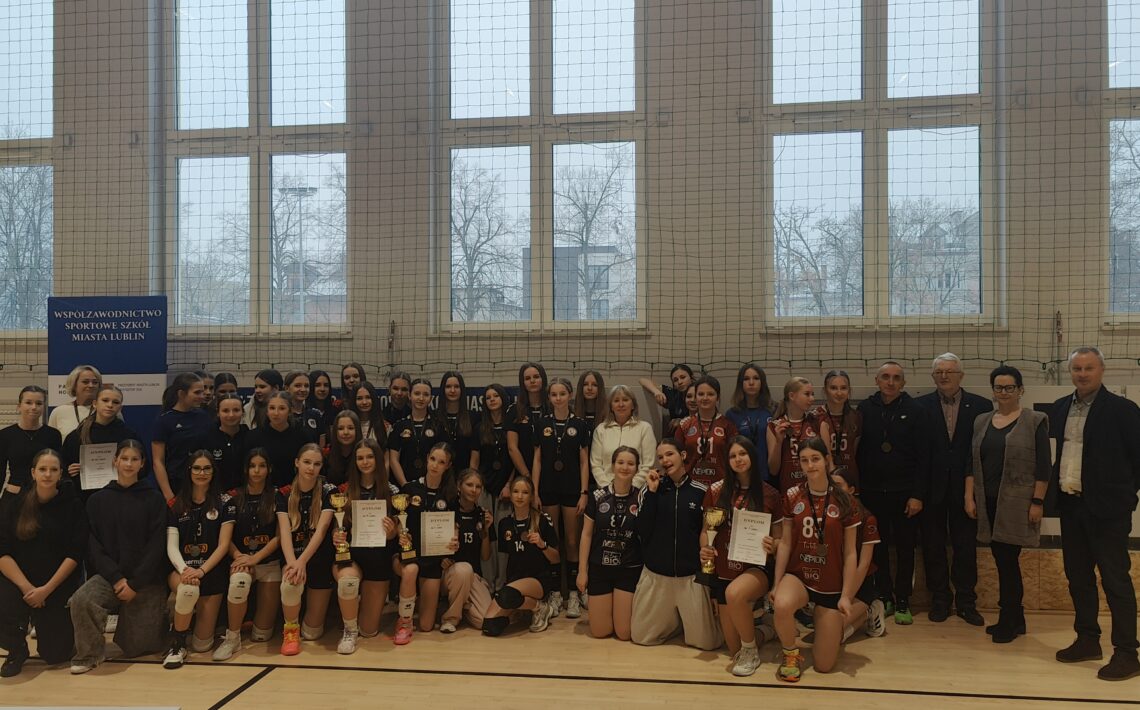 Zdjęcie grupowe uczestniczek turnieju siatkarskiego. Kilkadziesiąt dziewcząt w strojach sportowych, część z medalami i dyplomami. W tle baner informujący o współzawodnictwie sportowym szkół.