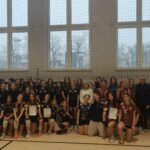 Zdjęcie grupowe uczestniczek turnieju siatkarskiego. Kilkadziesiąt dziewcząt w strojach sportowych, część z medalami i dyplomami. W tle baner informujący o współzawodnictwie sportowym szkół.