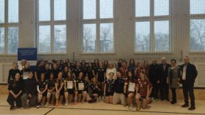 Zdjęcie grupowe uczestniczek turnieju siatkarskiego. Kilkadziesiąt dziewcząt w strojach sportowych, część z medalami i dyplomami. W tle baner informujący o współzawodnictwie sportowym szkół.