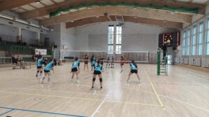 Mecz siatkówki dziewcząt w szkolnej hali sportowej. Dwie drużyny stoją po przeciwnych stronach siatki, zawodniczki w pozycjach obronnych. Po prawej stronie sędzia na podwyższeniu, w tle tablica wyników i trybuny.