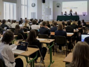 Szeroki widok sali obrad: wielu uczestników siedzi w ławkach z laptopami, z przodu na podwyższeniu panel prezydialny i osoba przemawiająca.