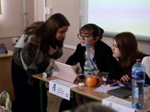 Trzy młode kobiety pracują wspólnie przy szkolnej ławce; jedna pochyla się i rozmawia, dwie siedzą i notują przy tablecie, na stole stoją butelki i materiały.