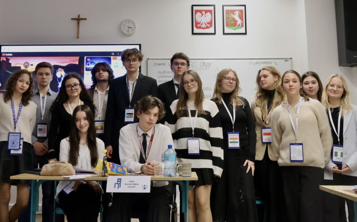 Duża grupa młodzieży pozuje do wspólnego zdjęcia w sali lekcyjnej; część osób stoi, część siedzi przy stole z materiałami i oznaczeniem funkcji.