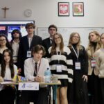 Duża grupa młodzieży pozuje do wspólnego zdjęcia w sali lekcyjnej; część osób stoi, część siedzi przy stole z materiałami i oznaczeniem funkcji.