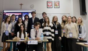 Duża grupa młodzieży pozuje do wspólnego zdjęcia w sali lekcyjnej; część osób stoi, część siedzi przy stole z materiałami i oznaczeniem funkcji.