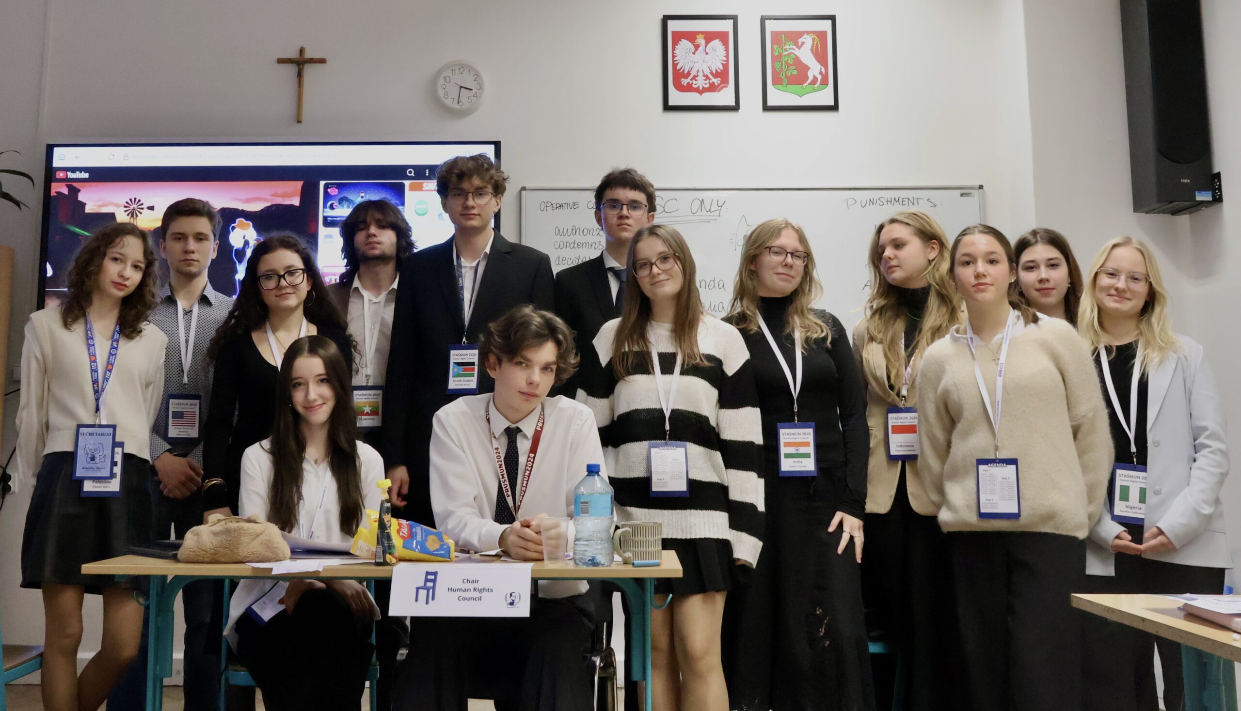 Duża grupa młodzieży pozuje do wspólnego zdjęcia w sali lekcyjnej; część osób stoi, część siedzi przy stole z materiałami i oznaczeniem funkcji.