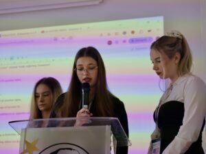 Trzy młode kobiety stoją przy mównicy; jedna czyta z kartki do mikrofonu, pozostałe słuchają, w tle widać wyświetloną prezentację.