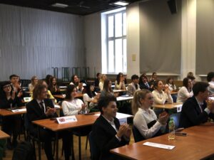 Uczestnicy zgromadzeni w sali wykładowej siedzą przy ławkach i klaszczą; na stołach widoczne są kartoniki z nazwami państw.