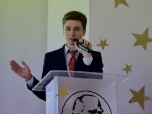 Młody mężczyzna w garniturze przemawia do mikrofonu zza przezroczystej mównicy, wykonując gest dłonią; tło jest jasne i oficjalne.