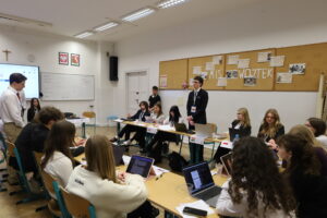 Uczestnicy formalnej debaty siedzą przy ustawionych w podkowę ławkach, korzystają z laptopów i notatek; jedna osoba stoi i zabiera głos.