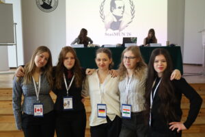 Grupa młodych kobiet stoi blisko siebie na scenie, obejmując się ramionami; wszystkie mają identyfikatory konferencyjne, w tle widoczny jest stół prezydialny.