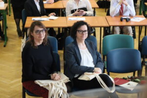 Dwie dorosłe kobiety siedzą w pierwszym rzędzie sali wykładowej, ubrane elegancko, trzymają jasne torby materiałowe; za nimi widać uczestników przy szkolnych ławkach.