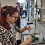 Uczennica związaną w kucyk fryzurą wykonuje miareczkowanie, trzymając zlewkę pod biuretą umocowaną na statywie. Na stanowisku znajduje się biała tryskawka, arkusz z notatkami oraz smartfon. W tle widać innych uczniów pracujących przy podobnych stanowiskach laboratoryjnych.