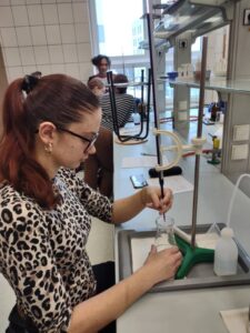 Uczennica związaną w kucyk fryzurą wykonuje miareczkowanie, trzymając zlewkę pod biuretą umocowaną na statywie. Na stanowisku znajduje się biała tryskawka, arkusz z notatkami oraz smartfon. W tle widać innych uczniów pracujących przy podobnych stanowiskach laboratoryjnych.