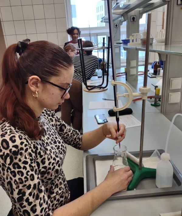 Uczennica związaną w kucyk fryzurą wykonuje miareczkowanie, trzymając zlewkę pod biuretą umocowaną na statywie. Na stanowisku znajduje się biała tryskawka, arkusz z notatkami oraz smartfon. W tle widać innych uczniów pracujących przy podobnych stanowiskach laboratoryjnych.