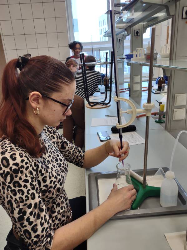 Uczennica związaną w kucyk fryzurą wykonuje miareczkowanie, trzymając zlewkę pod biuretą umocowaną na statywie. Na stanowisku znajduje się biała tryskawka, arkusz z notatkami oraz smartfon. W tle widać innych uczniów pracujących przy podobnych stanowiskach laboratoryjnych.