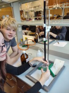 Dwie uczennice oraz uczeń pracują przy stanowisku do miareczkowania. Na pierwszym planie widoczna jest biureta zamocowana na statywie, z której do zlewki spływa roztwór. Na blacie stoją butelki z odczynnikami (brązowa butelka z etykietą), tryskawka z wodą destylowaną oraz palnik gazowy z pomarańczowym przewodem. Uczniowie koncentrują się na przebiegu doświadczenia w jasnym, nowoczesnym laboratorium.