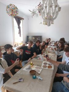 W jasnej sali muzealnej grupa młodzieży siedzi przy długim stole i bierze udział w warsztatach plastycznych. Uczniowie trzymają w dłoniach ozdobione jajka. Na stole znajdują się kartki z wzorami pisanek, świeczki, narzędzia do zdobienia i chusteczki. Nad stołem wiszą kolorowe, tradycyjne dekoracje z papieru. W tle stoi stare, ciemne pianino.