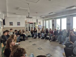 W jasnej szkolnej sali uczniowie siedzą na krzesłach ustawionych w dużym okręgu. W środku pozostawiono pustą przestrzeń. W spotkaniu bierze udział około dwudziestu kilku nastolatków oraz jedna dorosła kobieta. Większość osób siedzi zwrócona do środka kręgu i słucha rozmowy.

Sala jest dobrze oświetlona światłem dziennym wpadającym przez duże okna po prawej stronie. Na suficie znajduje się projektor multimedialny. Na ścianach wiszą plakaty i obrazy. W tle stoi przeszklona szafka z książkami i materiałami szkolnymi. Atmosfera przypomina zajęcia dyskusyjne lub warsztatowe.