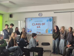 W klasie szkolnej uczniowie stoją i siedzą wokół dużego ekranu multimedialnego. Kilka osób stoi obok tablicy i prezentuje projekt lub prezentację. Pozostali uczniowie siedzą na krzesłach i słuchają.

Na ekranie widnieje slajd z napisem:

„CLASS OF 3A”

Nad napisem znajduje się tekst:
„I Liceum Ogólnokształcące im. Stanisława Staszica w Lublinie”

Pod napisem widnieją imiona:
Igor, Wiktoria, Norbert, Ola, Amelka, Kasia, Natalka

Slajd jest ozdobiony grafikami szkolnymi, takimi jak książki, zeszyty i przybory do nauki.