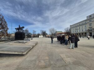 Na dużym miejskim placu stoi pomnik przedstawiający postać mężczyzny siedzącego na koniu. Pomnik znajduje się na wysokim, ciemnym cokole ustawionym na kamiennej platformie.

Po prawej stronie stoi grupa uczniów wraz z opiekunem. Słuchają osoby prowadzącej, która stoi kilka metrów przed nimi i coś tłumaczy. Wokół znajdują się budynki miejskie o jasnych elewacjach. Plac jest rozległy i wyłożony kamiennymi płytami. Na niebie widać rozległe, jasne chmury.