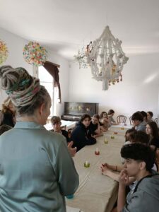 W tej samej sali warsztatowej starsza kobieta prowadzi zajęcia dla siedzącej przy stole młodzieży. Uczniowie słuchają i patrzą w jej stronę. Na stole stoją małe świeczki w kolorowych szklanych podstawkach. Nad stołem wiszą duże, tradycyjne ozdoby z papieru i bibuły.