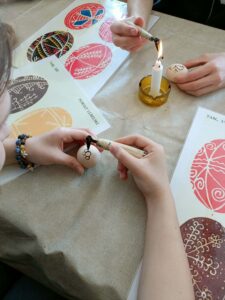 Zbliżenie na dłonie dwóch osób dekorujących jasne jajka. Jedna osoba rysuje wzór czarnym woskiem przy użyciu narzędzia podgrzanego nad płomieniem świecy. Na stole leżą plansze z kolorowymi wzorami pisanek oraz podpis „Powiat Lubelski” i oznaczenie „Tabl. XIII”.