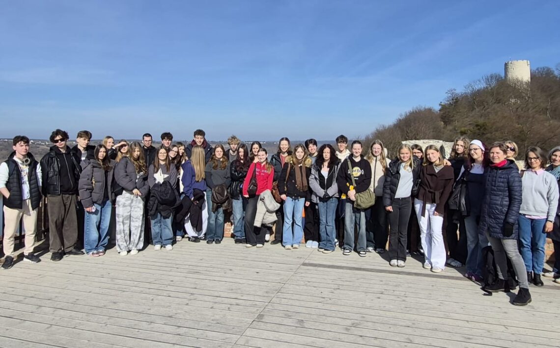 Duża grupa uczniów i kilku dorosłych stoi razem na drewnianym tarasie widokowym otoczonym niskim murem z czerwonej cegły. Wszyscy ustawieni są w jednym rzędzie i pozują do wspólnego zdjęcia. W tle rozciąga się szeroki krajobraz z doliną, drzewami i odległymi zabudowaniami. Po prawej stronie na wzgórzu widać kamienną wieżę ruin zamku. Pogoda jest słoneczna, a niebo jasnoniebieskie.
