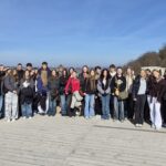 Duża grupa uczniów i kilku dorosłych stoi razem na drewnianym tarasie widokowym otoczonym niskim murem z czerwonej cegły. Wszyscy ustawieni są w jednym rzędzie i pozują do wspólnego zdjęcia. W tle rozciąga się szeroki krajobraz z doliną, drzewami i odległymi zabudowaniami. Po prawej stronie na wzgórzu widać kamienną wieżę ruin zamku. Pogoda jest słoneczna, a niebo jasnoniebieskie.