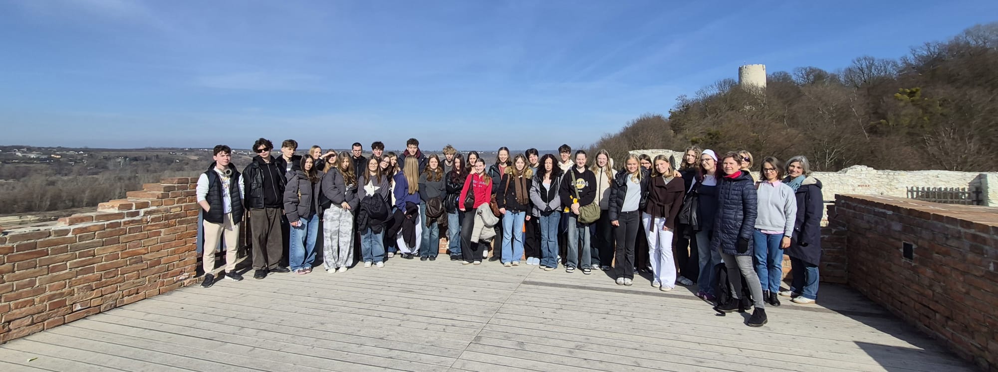Duża grupa uczniów i kilku dorosłych stoi razem na drewnianym tarasie widokowym otoczonym niskim murem z czerwonej cegły. Wszyscy ustawieni są w jednym rzędzie i pozują do wspólnego zdjęcia. W tle rozciąga się szeroki krajobraz z doliną, drzewami i odległymi zabudowaniami. Po prawej stronie na wzgórzu widać kamienną wieżę ruin zamku. Pogoda jest słoneczna, a niebo jasnoniebieskie.