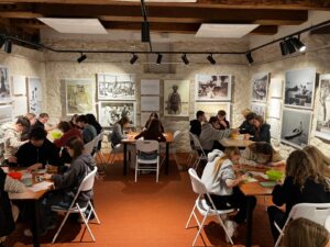 W pomieszczeniu o kamiennych ścianach, przypominającym muzeum lub salę warsztatową, kilka grup uczniów siedzi przy stołach. Każda grupa pracuje nad zadaniem plastycznym lub rękodzielniczym.

Na stołach leżą kartki papieru, narzędzia i miski z materiałami. Uczniowie pochylają się nad swoją pracą. Na ścianach wiszą duże czarno-białe fotografie przedstawiające ludzi i sceny historyczne. Nad stołami zamontowane są lampy punktowe oświetlające przestrzeń.