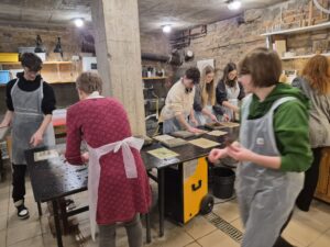 W warsztatowej pracowni o surowych, ceglastych ścianach grupa uczniów pracuje przy długim stole roboczym. Wszyscy mają na sobie ochronne plastikowe fartuchy.

Na stole znajdują się arkusze papieru, ramki i narzędzia używane do pracy ręcznej, prawdopodobnie przy tworzeniu papieru lub grafik. Uczniowie wykonują różne czynności – układają arkusze, dociskają je lub przygotowują materiały.

Pomieszczenie oświetlają lampy sufitowe. W tle widać półki z narzędziami oraz zlew do pracy warsztatowej.