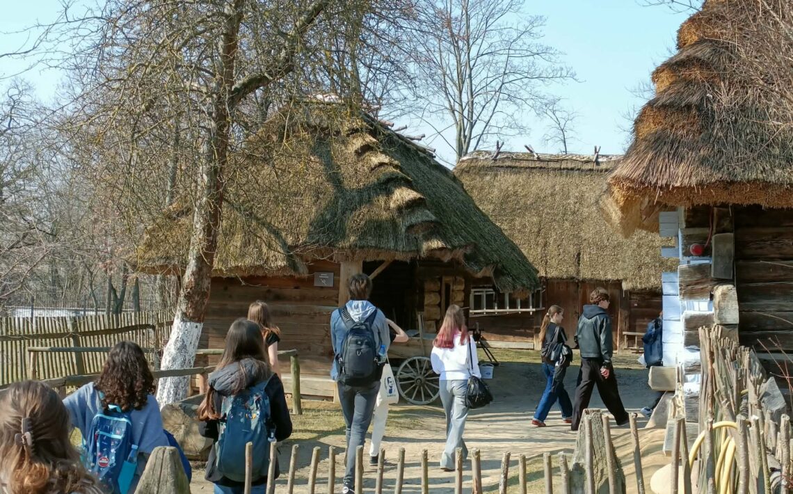 Grupa młodzieży spaceruje po terenie skansenu między drewnianymi chatami krytymi strzechą. Wokół znajdują się drewniane płoty, drzewa i elementy dawnej zabudowy wiejskiej, takie jak wóz. Uczniowie idą ścieżką z plecakami, zwiedzając teren muzeum na świeżym powietrzu.
