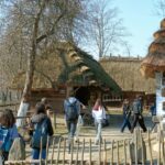 Grupa młodzieży spaceruje po terenie skansenu między drewnianymi chatami krytymi strzechą. Wokół znajdują się drewniane płoty, drzewa i elementy dawnej zabudowy wiejskiej, takie jak wóz. Uczniowie idą ścieżką z plecakami, zwiedzając teren muzeum na świeżym powietrzu.