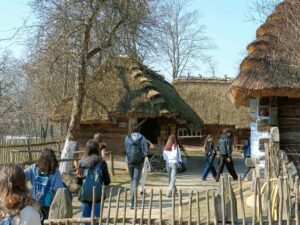 Grupa młodzieży spaceruje po terenie skansenu między drewnianymi chatami krytymi strzechą. Wokół znajdują się drewniane płoty, drzewa i elementy dawnej zabudowy wiejskiej, takie jak wóz. Uczniowie idą ścieżką z plecakami, zwiedzając teren muzeum na świeżym powietrzu.