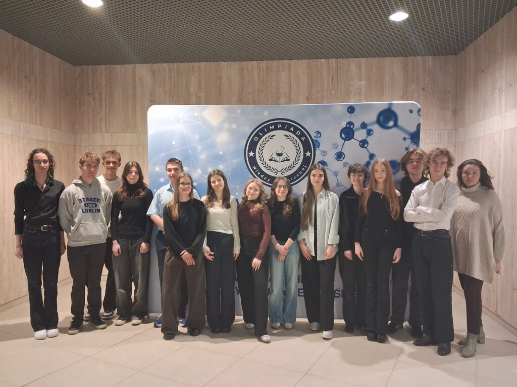 Grupa kilkunastu uczniów wraz z opiekunką stoi w rzędzie we wnętrzu budynku o jasnych, drewnianych ścianach. Za nimi znajduje się duża tablica z napisem „Olimpiada Wiedzy Chemicznej” oraz grafiką cząsteczek. Uczestnicy są ubrani w stroje półformalne (koszule, swetry, spodnie), stoją blisko siebie i patrzą w stronę aparatu. Atmosfera zdjęcia jest oficjalna i reprezentacyjna.