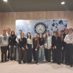 Grupa kilkunastu uczniów wraz z opiekunką stoi w rzędzie we wnętrzu budynku o jasnych, drewnianych ścianach. Za nimi znajduje się duża tablica z napisem „Olimpiada Wiedzy Chemicznej” oraz grafiką cząsteczek. Uczestnicy są ubrani w stroje półformalne (koszule, swetry, spodnie), stoją blisko siebie i patrzą w stronę aparatu. Atmosfera zdjęcia jest oficjalna i reprezentacyjna.