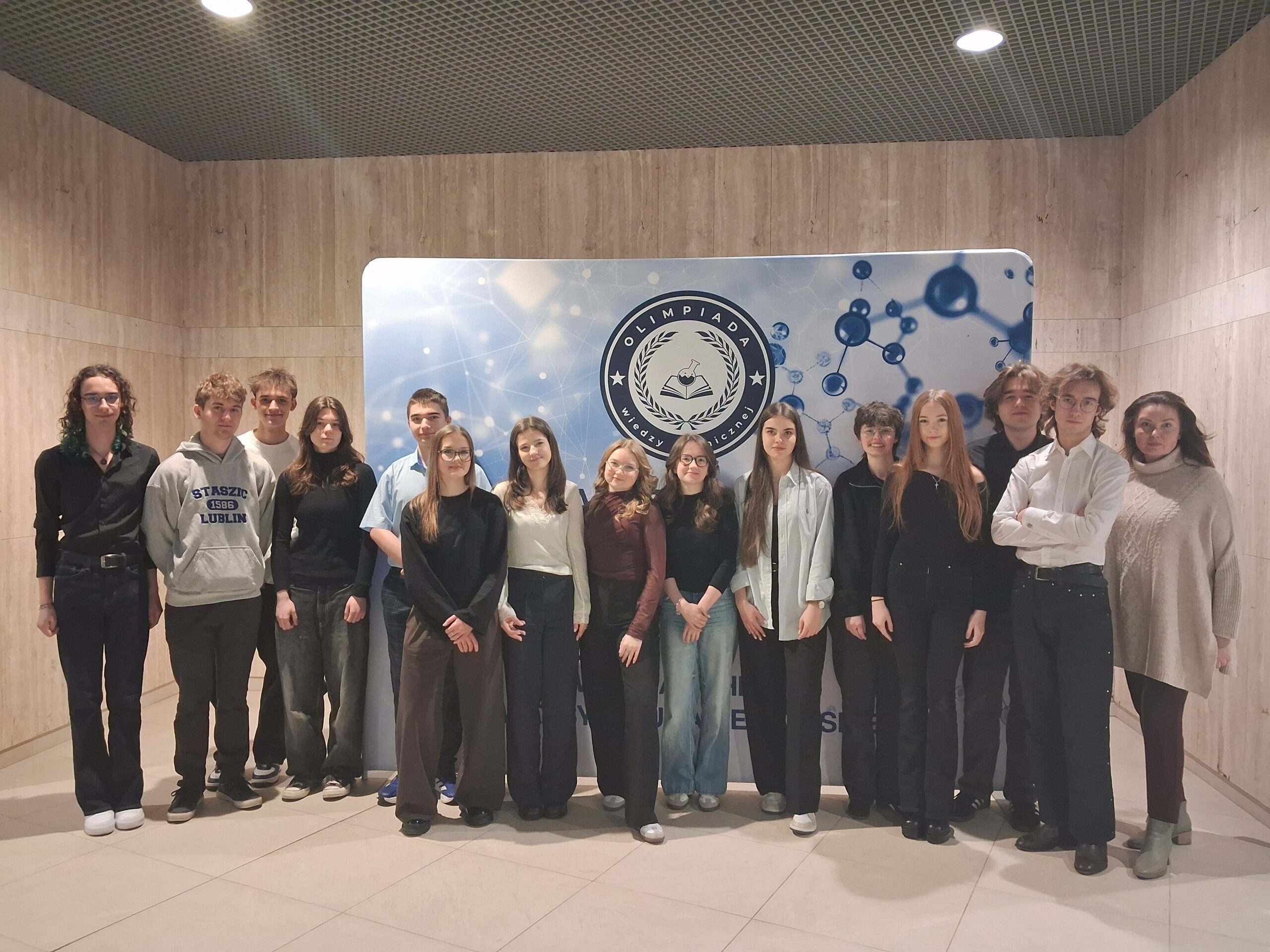 Grupa kilkunastu uczniów wraz z opiekunką stoi w rzędzie we wnętrzu budynku o jasnych, drewnianych ścianach. Za nimi znajduje się duża tablica z napisem „Olimpiada Wiedzy Chemicznej” oraz grafiką cząsteczek. Uczestnicy są ubrani w stroje półformalne (koszule, swetry, spodnie), stoją blisko siebie i patrzą w stronę aparatu. Atmosfera zdjęcia jest oficjalna i reprezentacyjna.