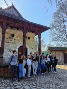 Grupa kilkunastu nastolatków stoi na brukowanym placu przed jasnym, kamiennym budynkiem z drewnianym gankiem i kolumnami. Nad drzwiami widnieje napis „Muzeum Wsi Lubelskiej” oraz łukowaty napis „WEJŚCIE”. W tle widać bezlistne drzewa i niebieskie niebo. Uczniowie stoją obok siebie, część z plecakami i kurtkami, jak do wspólnego zdjęcia podczas wycieczki.