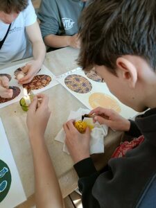 Zbliżenie na uczniów podczas zdobienia jajka metodą woskową. Jeden z chłopców trzyma w dłoni żółte jajko i rysuje na nim wzór rozgrzanym narzędziem z woskiem. Obok płonie mała świeczka służąca do podgrzewania narzędzia. Na stole leżą kartki z przykładami tradycyjnych wzorów pisanek.