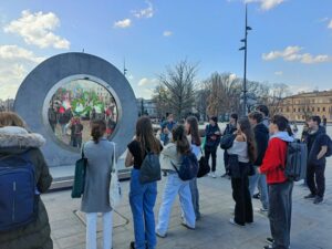 Grupa młodzieży stoi na miejskim placu i patrzy na dużą, okrągłą instalację przypominającą betonową ramę z ekranem w środku. Na ekranie wyświetlany jest obraz ludzi oraz zielony symbol przypominający koniczynę. Uczniowie stoją półkolem, część z plecakami, słuchając lub obserwując instalację. W tle widać drzewa, budynki miasta i latarnie uliczne.