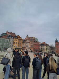 Grupa uczniów z nauczycielką stoi na Rynku Starego Miasta w Warszawie na tle historycznych kamienic.