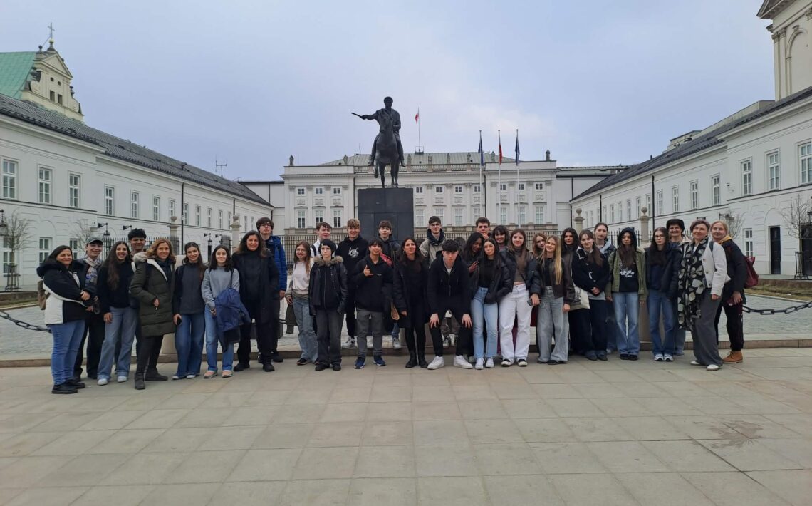 Grupa uczniów i nauczycieli stoi przed pomnikiem na koniu na placu przed Pałacem Prezydenckim w Warszawie.