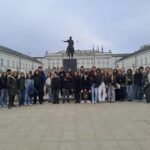 Grupa uczniów i nauczycieli stoi przed pomnikiem na koniu na placu przed Pałacem Prezydenckim w Warszawie.