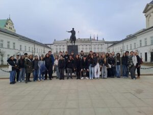 Grupa uczniów i nauczycieli stoi przed pomnikiem na koniu na placu przed Pałacem Prezydenckim w Warszawie.