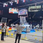 Ponownie widok hali zawodów robotycznych FIRST z boku pola gry. Na planszy rozrzucone są liczne żółte piłki. Kilka osób w szarych koszulkach obserwuje lub steruje robotami. W tle widać trybuny oraz duże ekrany z transmisją z pola gry.