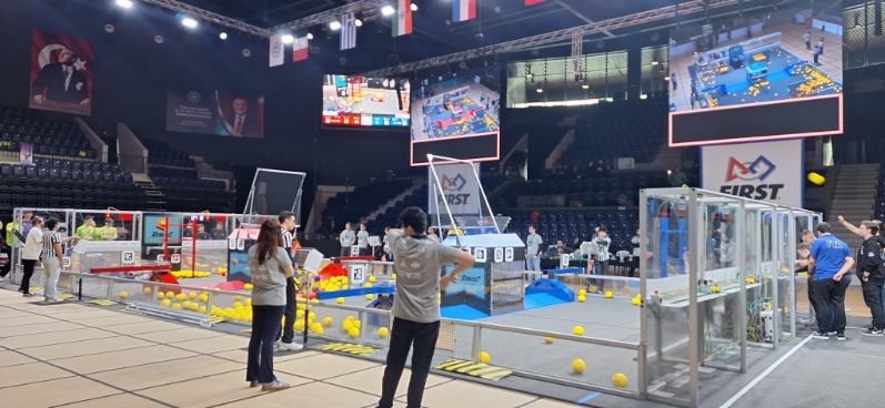 Ponownie widok hali zawodów robotycznych FIRST z boku pola gry. Na planszy rozrzucone są liczne żółte piłki. Kilka osób w szarych koszulkach obserwuje lub steruje robotami. W tle widać trybuny oraz duże ekrany z transmisją z pola gry.