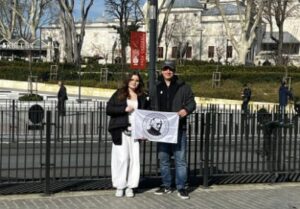 Na zewnątrz, przed ogrodzeniem i historycznym budynkiem, stoją dwie osoby – kobieta i mężczyzna. Trzymają rozłożoną flagę z czarno-białą grafiką przedstawiającą portret osoby w okręgu. Za nimi widać drzewa bez liści i jasny budynek przypominający obiekt zabytkowy.