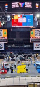 Widok z góry na halę zawodów robotycznych. W centrum znajduje się pole gry z dużą liczbą żółtych piłek ułożonych w stos. Wokół pracują uczestnicy i sędziowie przygotowujący elementy gry. Nad areną widoczny jest ogromny ekran z wynikami oraz napis „Fikret Yüksel Foundation”.