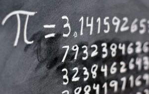 Na zdjęciu widać fragment tablicy lub ciemnego tła, na którym białą kredą zapisano symbol matematyczny liczby π (pi) oraz jej rozwinięcie dziesiętne zaczynające się od 3,141592653…. Liczby zapisane są w kilku liniach, a część zapisu jest lekko rozmazana, jakby ktoś próbował zetrzeć fragment kredy. Obraz przedstawia motyw matematyczny związany z liczbą pi